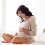 A pregnant person sits cross-legged on a bed, gently cradling their belly in a calm, sunlit room—representing peaceful anticipation, self-connection, and holistic prenatal wellness.
