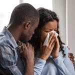 A woman wipes away tears with a tissue while being comforted by a supportive partner—capturing a moment of emotional vulnerability, empathy, and the importance of compassionate connection during times of grief or distress.
