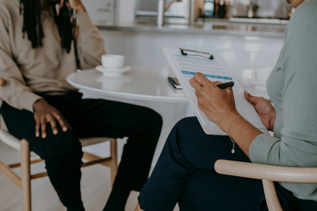 A therapist takes notes on a clipboard during a one-on-one session with a client, seated at a small table in a calm, welcoming environment—representing compassionate care and professional support.