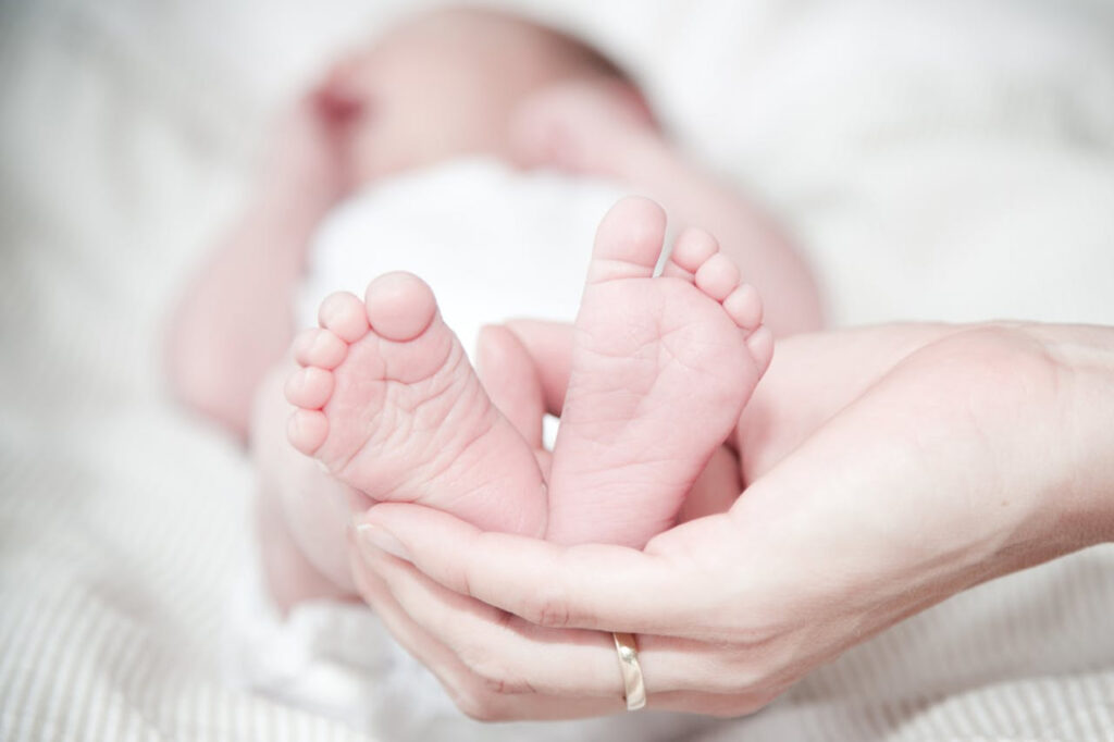 A caregiver gently cradles the feet of a newborn baby lying on a soft surface—symbolizing tenderness, early bonding, and the nurturing support of infant and postpartum care.