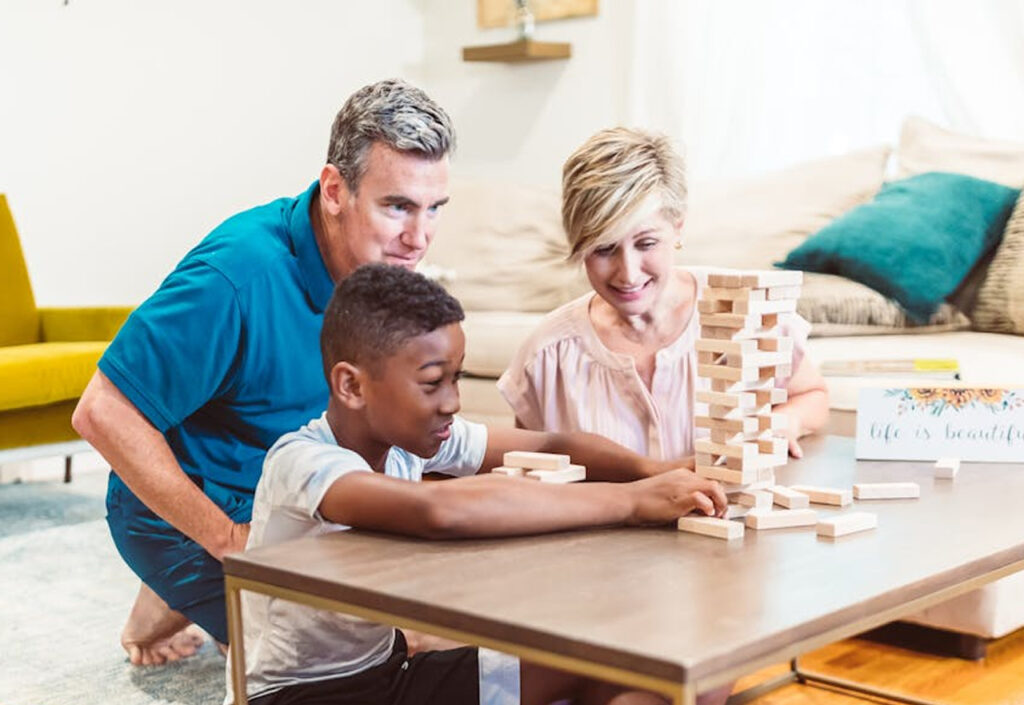 A child plays a game of Jenga with two adults at a low table, all smiling and engaged—illustrating playful connection, developmental growth, and supportive caregiving in a home-like environment.