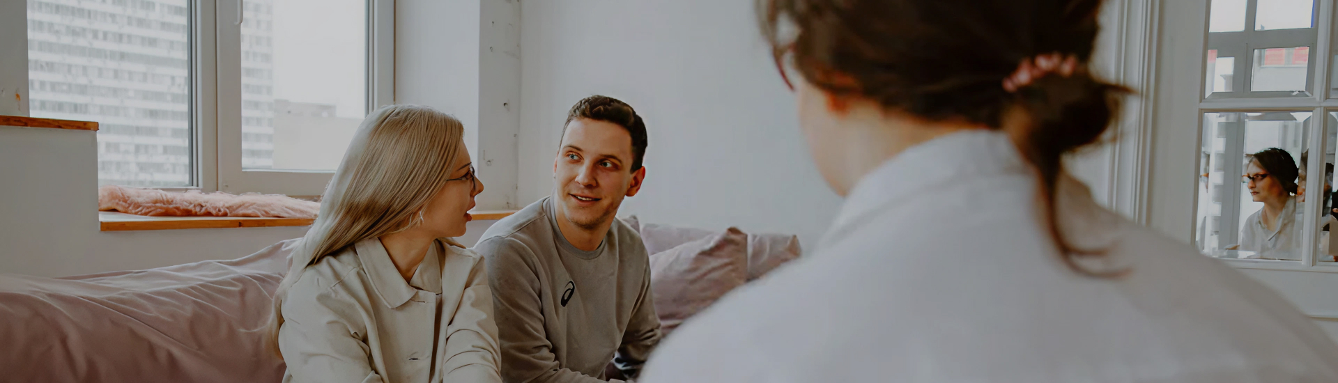 A couple sits on a couch speaking with a healthcare professional in a light-filled room—representing supportive consultation, relationship-centered care, and open dialogue in a safe therapeutic space.