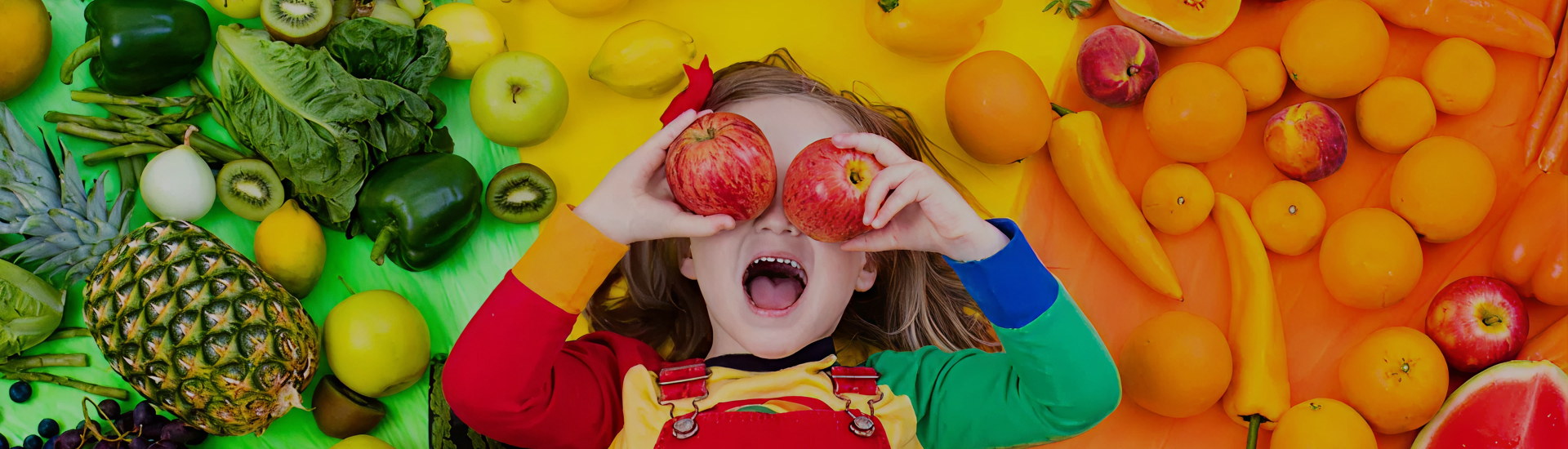 A joyful child wearing a rainbow-colored outfit holds apples over their eyes, surrounded by a vibrant rainbow of fruits and vegetables—celebrating colorful, healthy eating and making nutrition fun for kids.