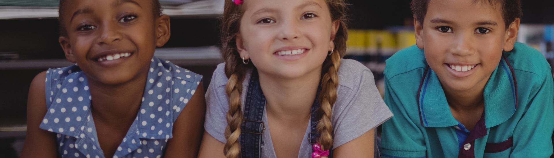Three smiling children sit close together in a classroom or learning space, representing joy, diversity, and the importance of early childhood development and inclusive care.