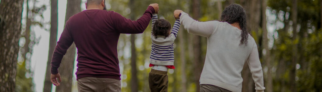 A family walks hand in hand through a forest trail, with a child playfully lifted between two adults—symbolizing connection, support, and the joy of shared outdoor moments. And emphasizing the need for parents therapy