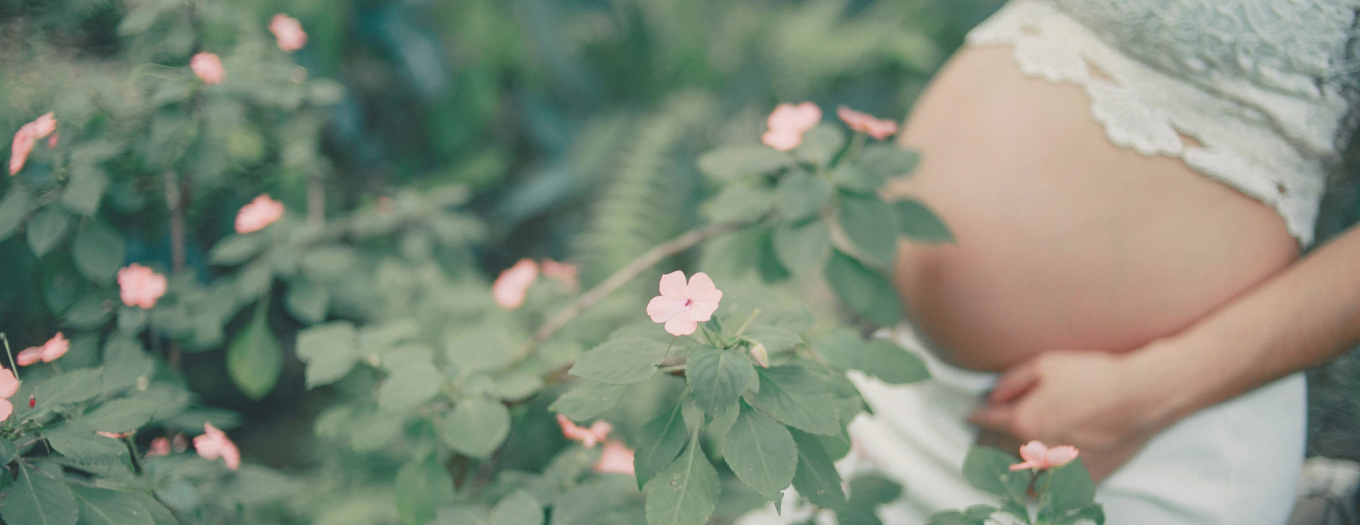 A pregnant person gently holding their bare belly while surrounded by soft pink flowers and greenery, symbolizing natural growth, nurturing, and perinatal wellness.