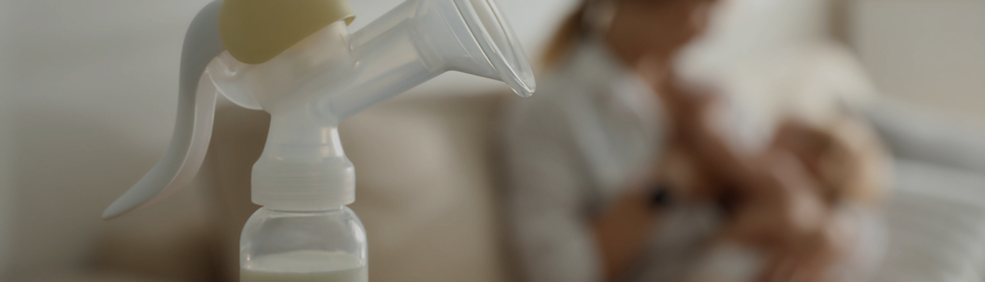 A breast pump sits in focus on a table while a parent holds and feeds their baby in the softly blurred background—representing the balance of breastfeeding, pumping, and nurturing infant care.