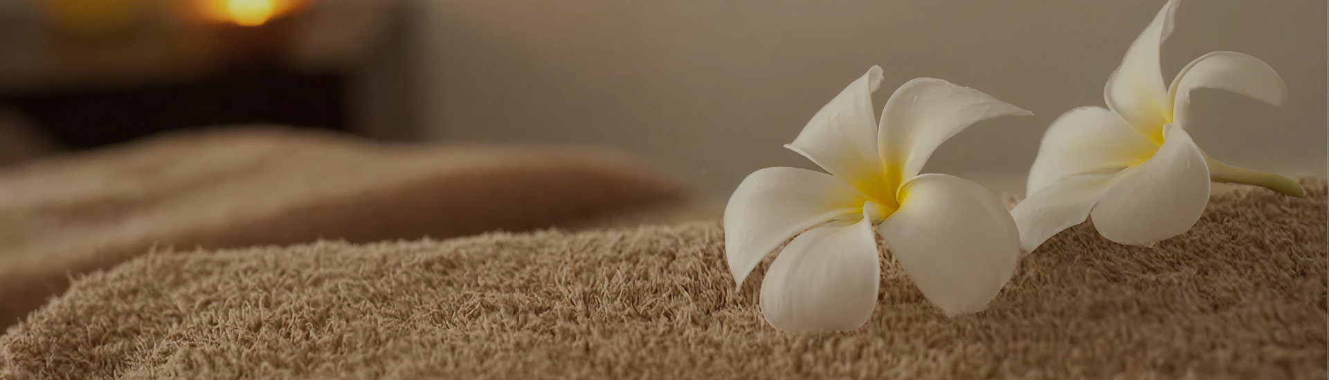 Two white plumeria flowers rest on a folded towel in a softly lit space—evoking calm, balance, and the restorative environment of therapeutic massage or holistic wellness treatments.