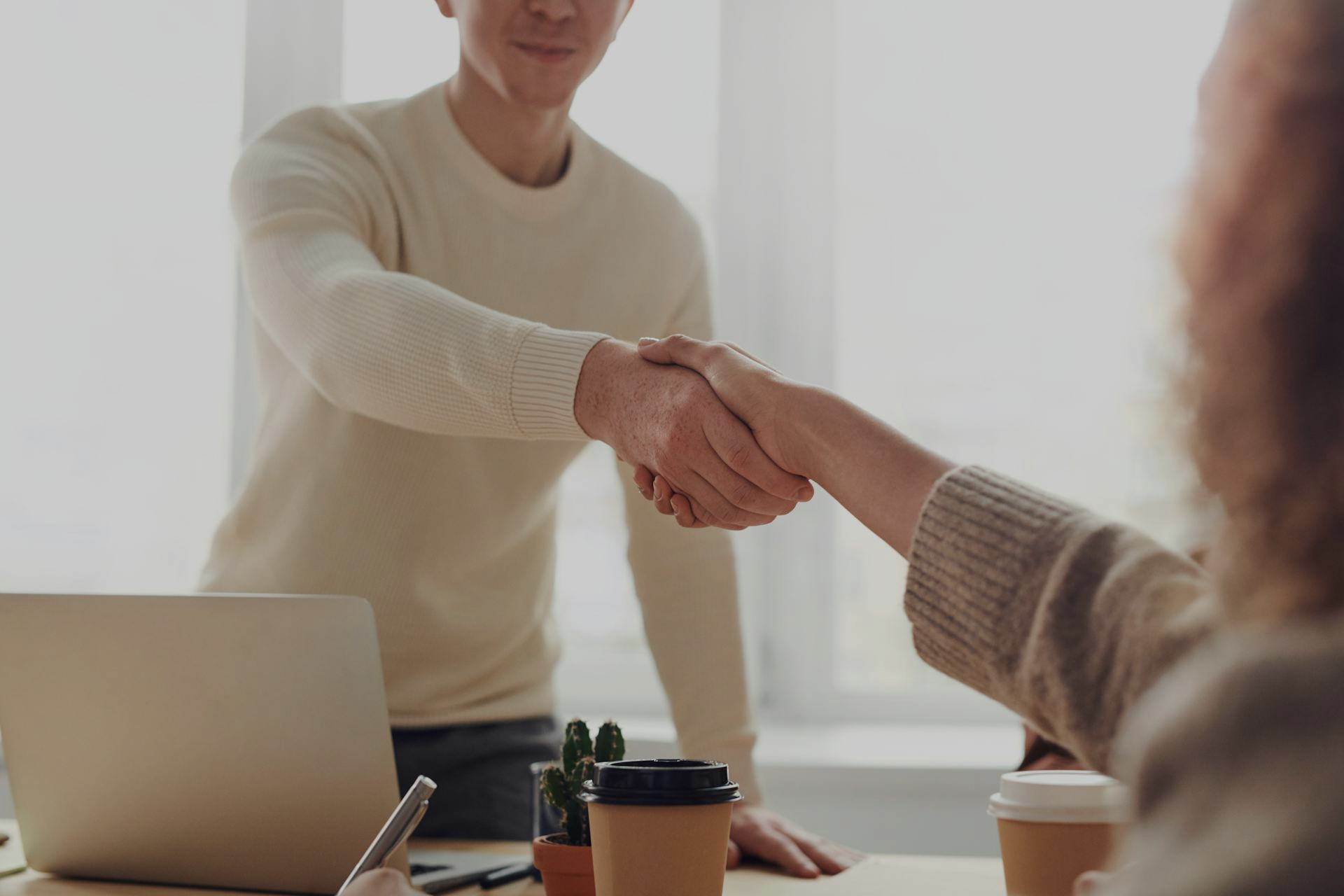 Two people shaking hands across a table in a bright, welcoming space—representing collaboration, support, and the beginning of a trusted therapeutic or professional relationship.
