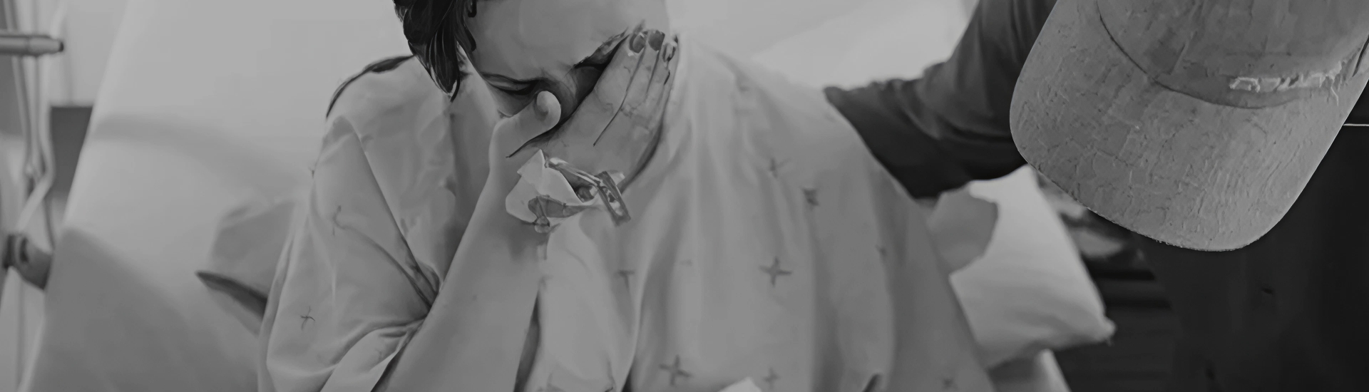 In a black-and-white image, a person in a hospital gown sits with their face in their hand, visibly overwhelmed, while another offers a comforting hand on their back—symbolizing grief, emotional pain, and the importance of supportive presence during vulnerable moments.