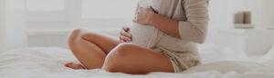 A pregnant person sits cross-legged on a bed, gently cradling their belly in a calm, sunlit room—representing peaceful anticipation, self-connection, and holistic prenatal wellness.