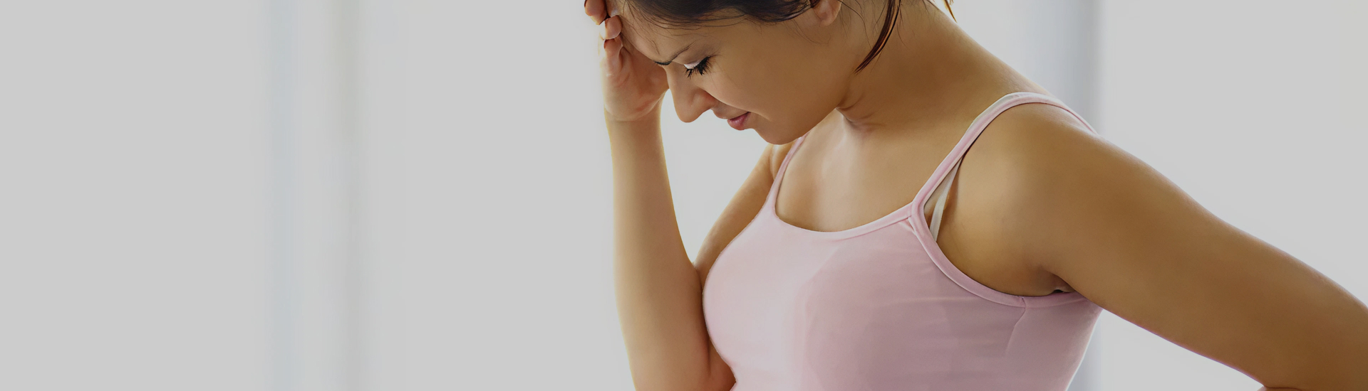 A pregnant woman in a pink tank top stands with her hand on her forehead, appearing stressed or deep in thought.