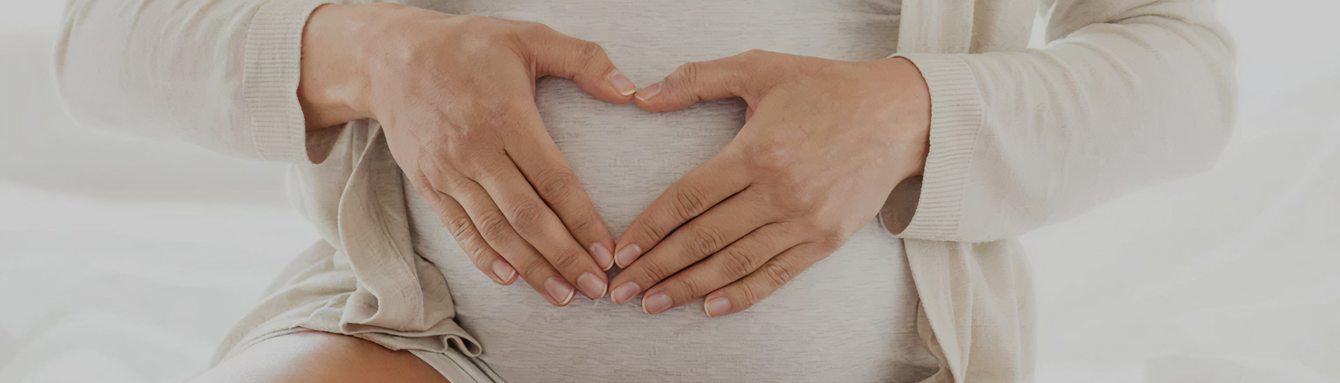 Close-up of a pregnant person’s hands gently forming a heart shape over their belly, sitting on a light-colored bed while wearing a soft beige cardigan.