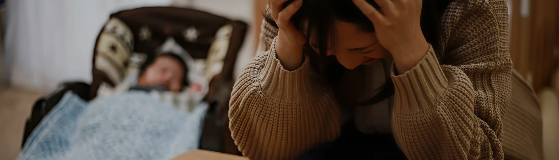 A distressed parent sits with their head in their hands while a baby rests in the background—capturing the emotional weight of postpartum overwhelm and the need for support in early parenthood.