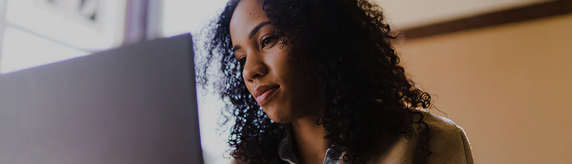 A focused woman sits at a laptop in a softly lit room, suggesting remote access to care, virtual therapy, or self-guided wellness from the comfort of home