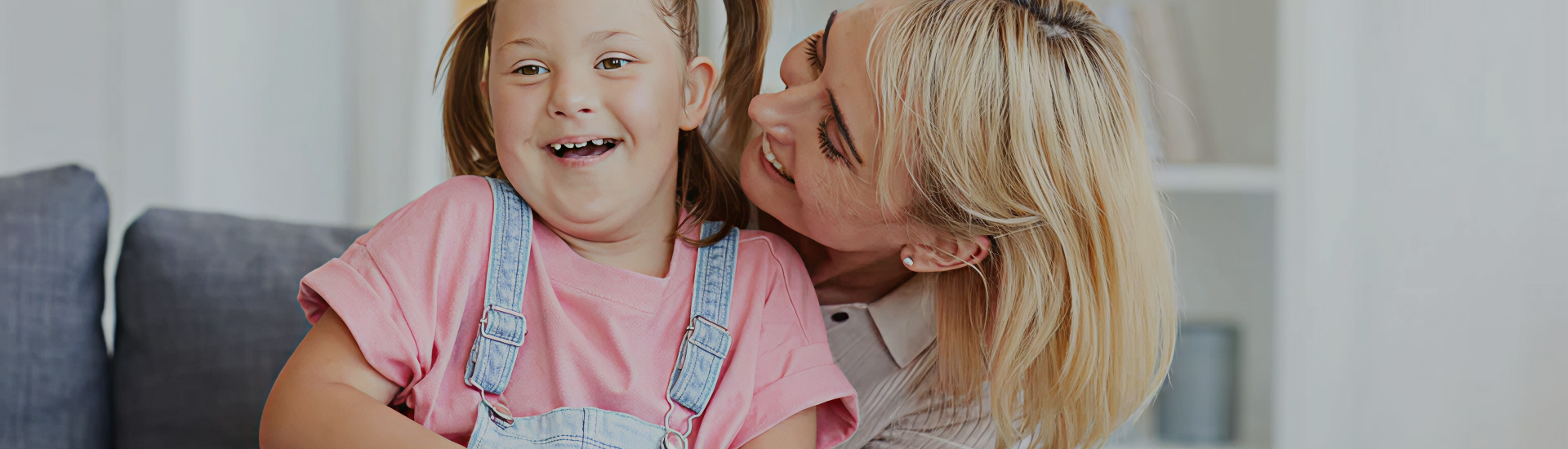 A smiling child with Down syndrome is embraced by a caregiver on a couch, both sharing a joyful moment—highlighting unconditional love, support, and inclusive family wellness.