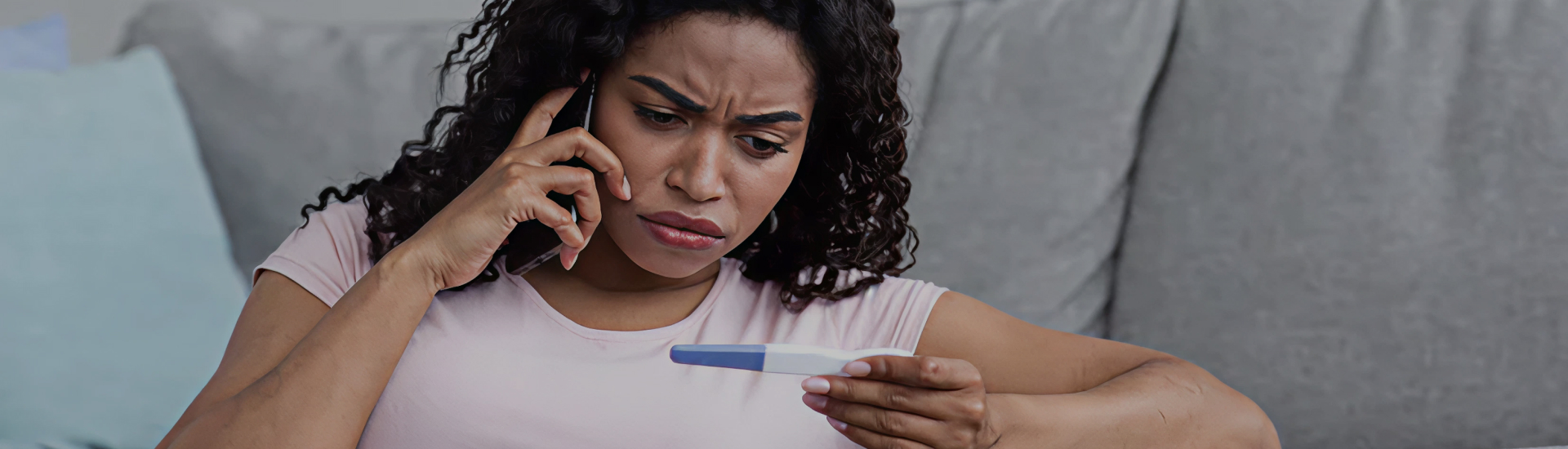 A woman sitting on a couch looks at a pregnancy test with concern while speaking on the phone.