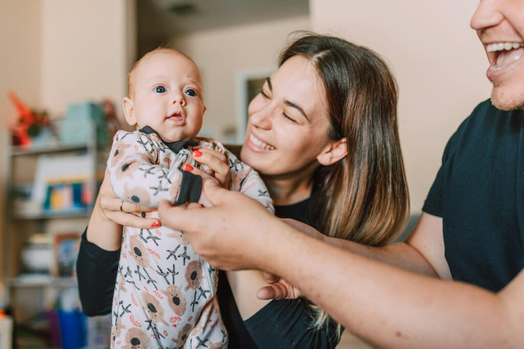 Two smiling adults engage playfully with their baby, who is dressed in a patterned onesie—capturing warmth, early bonding, and the joyful moments of family connection.