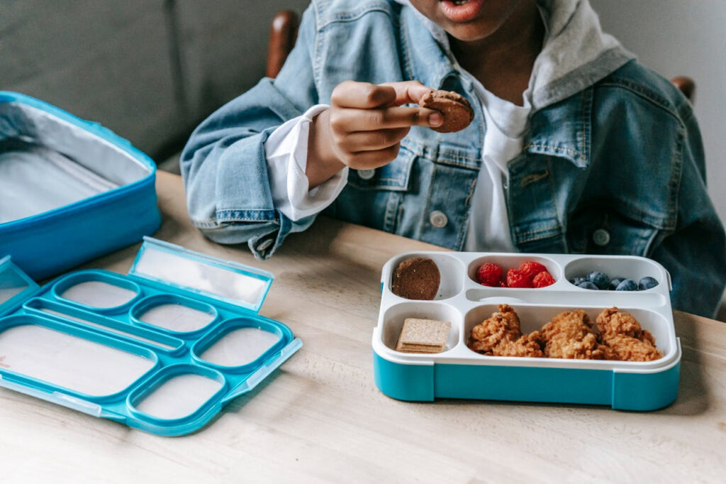A child eats from a balanced lunchbox filled with fruits, protein, and snacks, highlighting the role of nutritious meals in supporting childhood wellness, focus, and healthy development.