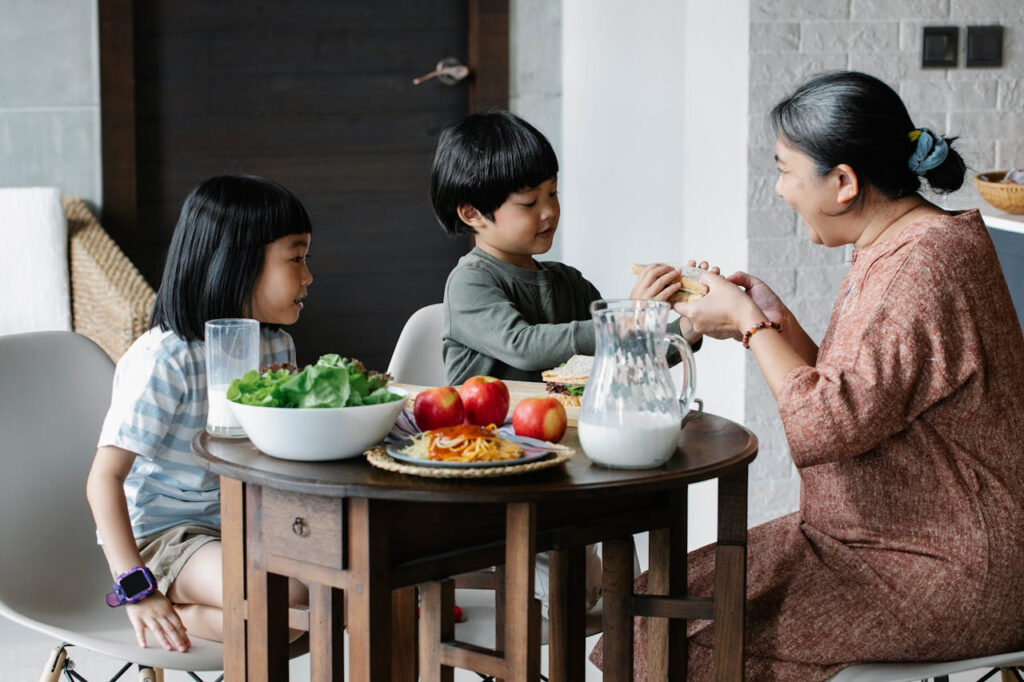 An adult and two children sit together at a dining table, sharing a nutritious meal with fresh fruits, vegetables, and milk—highlighting the importance of family connection, nourishment, and healthy eating habits.