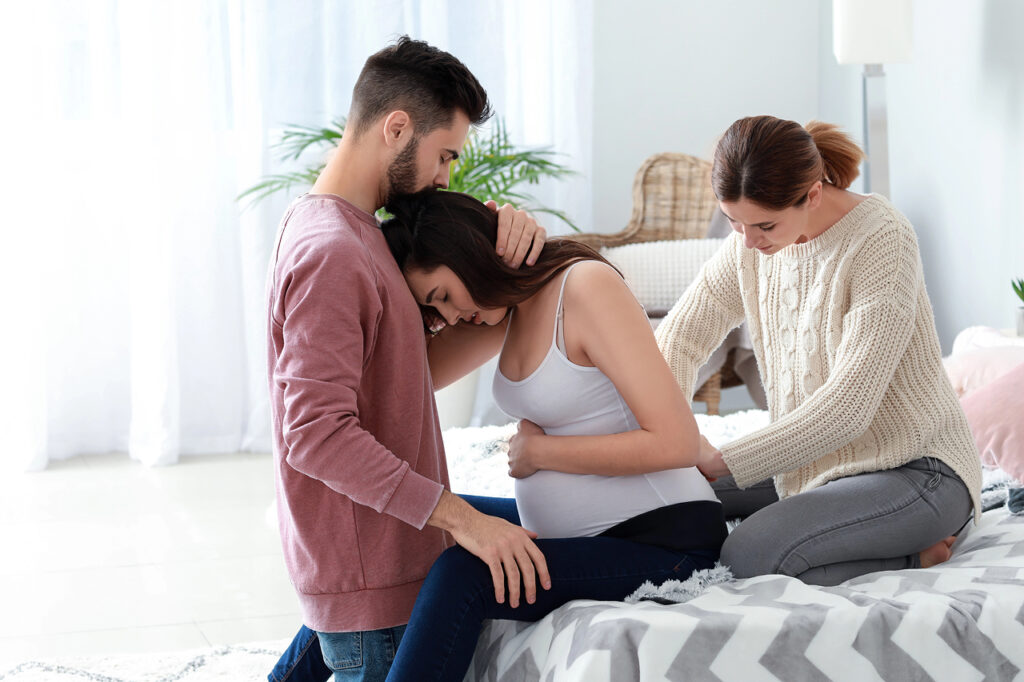 A pregnant person leans into their partner for support while a birth professional or doula provides physical comfort from behind—capturing a moment of connection, care, and continuous support during labor.
