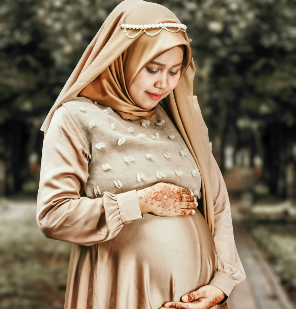 A pregnant woman wearing a light brown hijab and a long dress stands outdoors on a path, gently cradling her belly with both hands. She looks down lovingly with a peaceful smile, surrounded by soft-focus trees in the background.