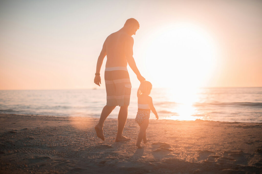 A parent and child walk hand in hand along the beach at sunset, silhouetted by the glowing sun—symbolizing presence, bonding, and the grounding power of nature and family connection.