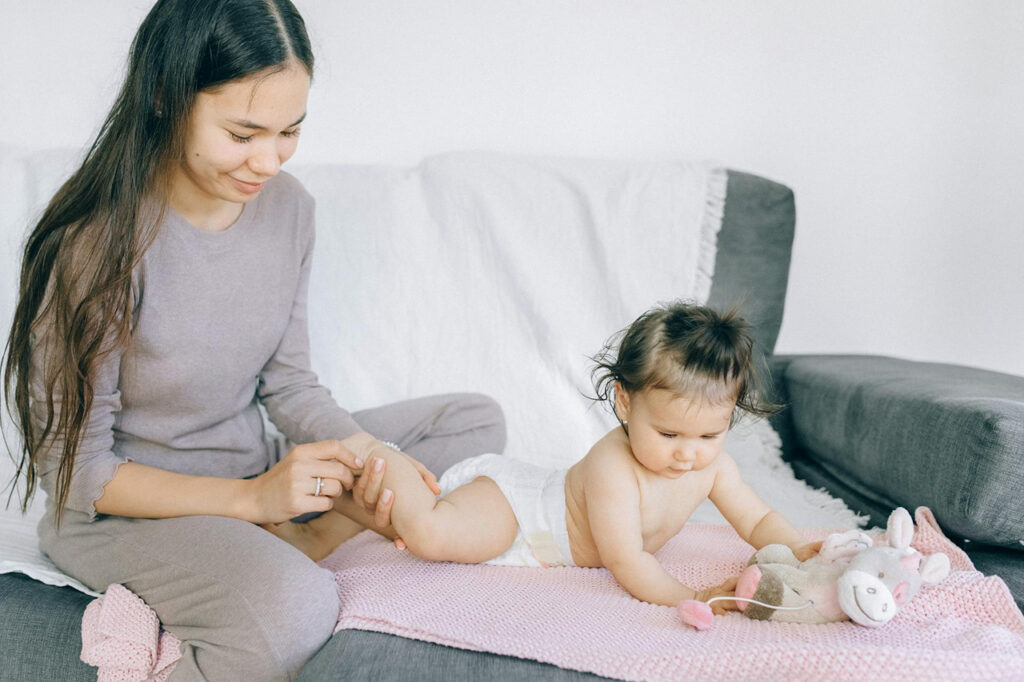 A parent gently massages their baby’s leg while the child plays with a stuffed toy on a soft blanket—illustrating loving touch, bonding, and holistic care during early development.
