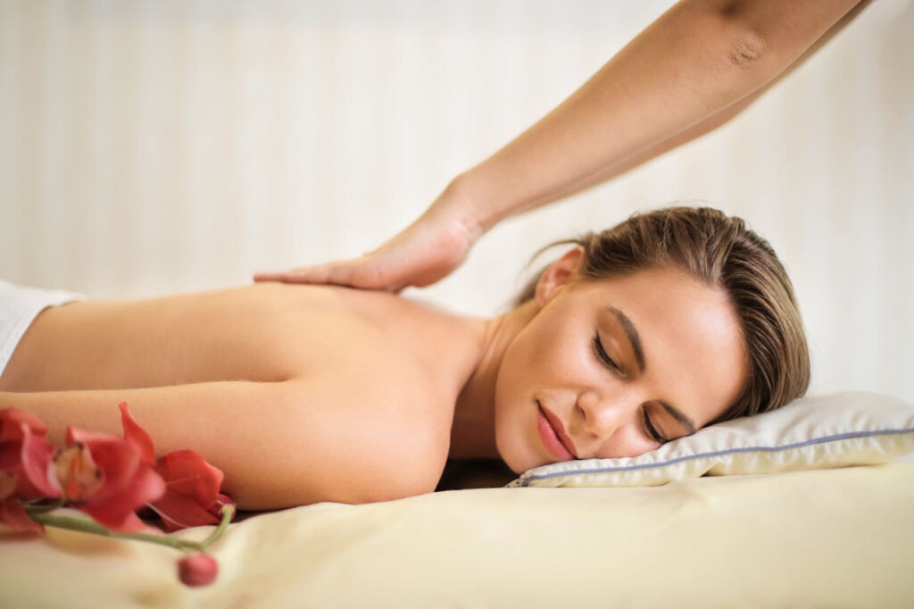 A woman lies peacefully on a massage table with her eyes closed as a therapist applies gentle pressure to her back—symbolizing relaxation, stress relief, and the restorative benefits of therapeutic massage.
