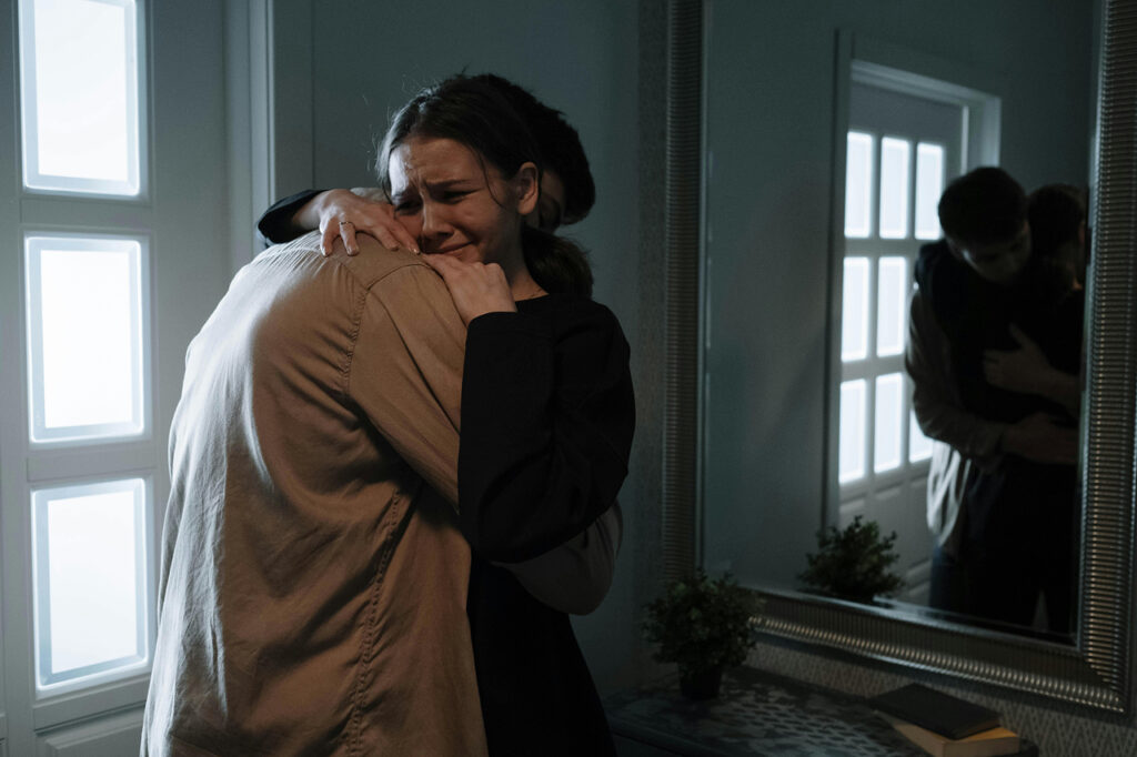 Two people embrace tightly in a dimly lit room, one visibly emotional with tears—capturing a moment of deep grief, comfort, and the power of human connection during times of distress.