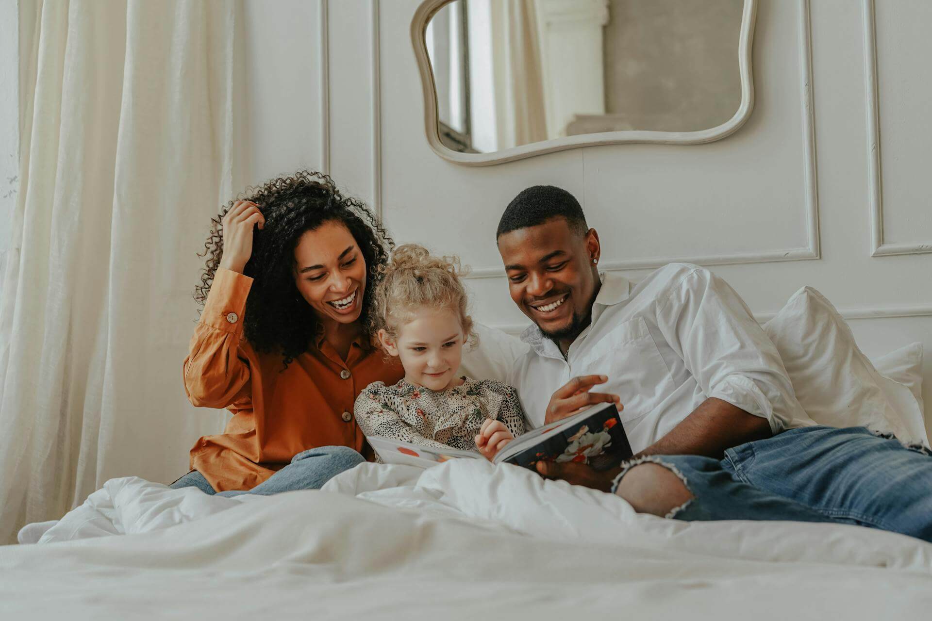 A joyful family of three sits together on a bed, smiling and reading a book, reflecting love, connection, and nurturing moments in early childhood and family life.