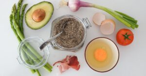 A variety of fresh, whole foods—including avocado, asparagus, tomato, egg, chia seeds, green onion, and prosciutto—arranged on a white surface, representing balanced nutrition and mindful eating.