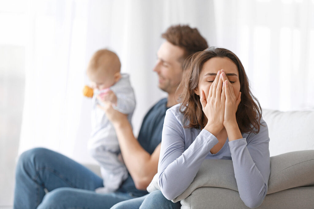 A visibly overwhelmed mother sits on a couch with her hands covering her face, while a father in the background holds their baby. The scene reflects emotional exhaustion, parental stress, or postpartum challenges.
