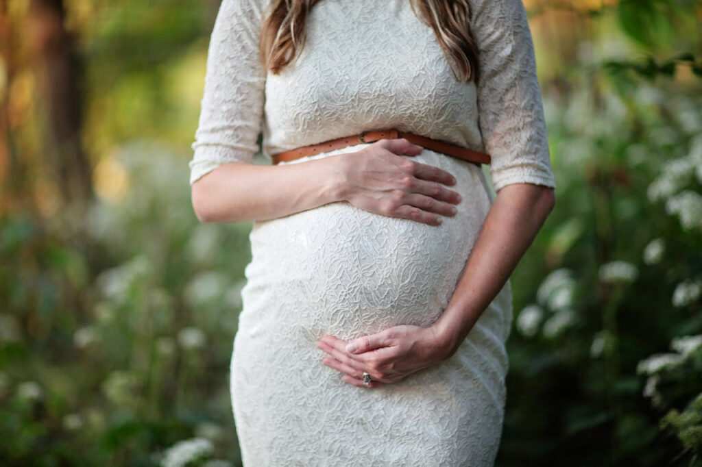 Pregnant woman standing outdoors in a garden setting, wearing a white lace dress with a brown belt. Her hands are gently placed on her belly, surrounded by soft greenery and white flowers