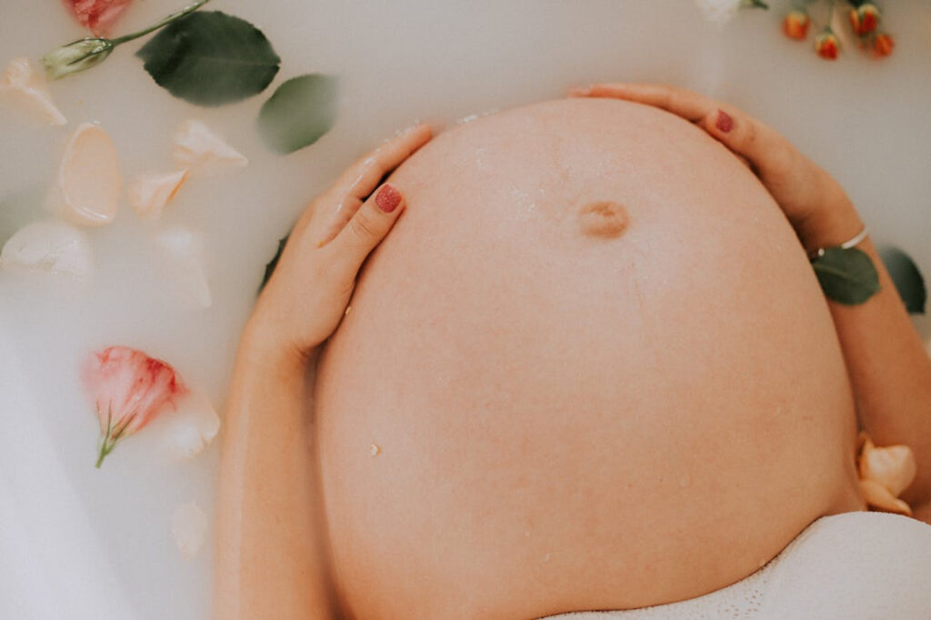 Close-up of a pregnant belly in a milk bath surrounded by floating flowers and petals. The person’s hands gently rest on their stomach, creating a serene and intimate atmosphere.