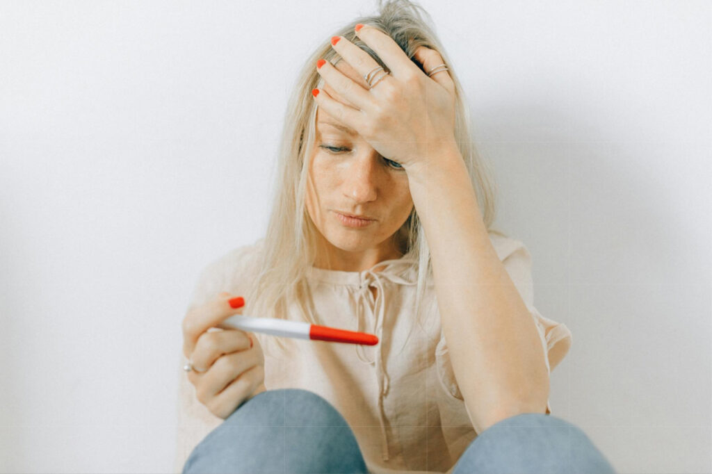 Woman with hand on forehead holding a pregnancy test looking disappointed symbolizing infertility or problems conceiving