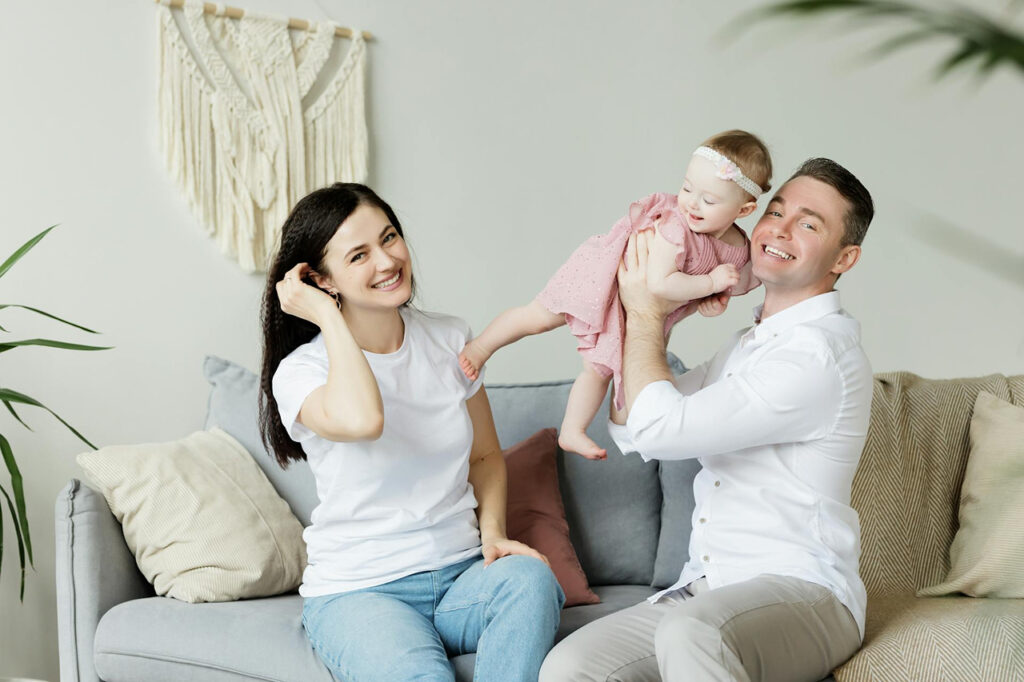 A cheerful family sits together on a couch, with one parent lifting their smiling baby into the air while the other looks on—capturing love, laughter, and connection in early parenthood.