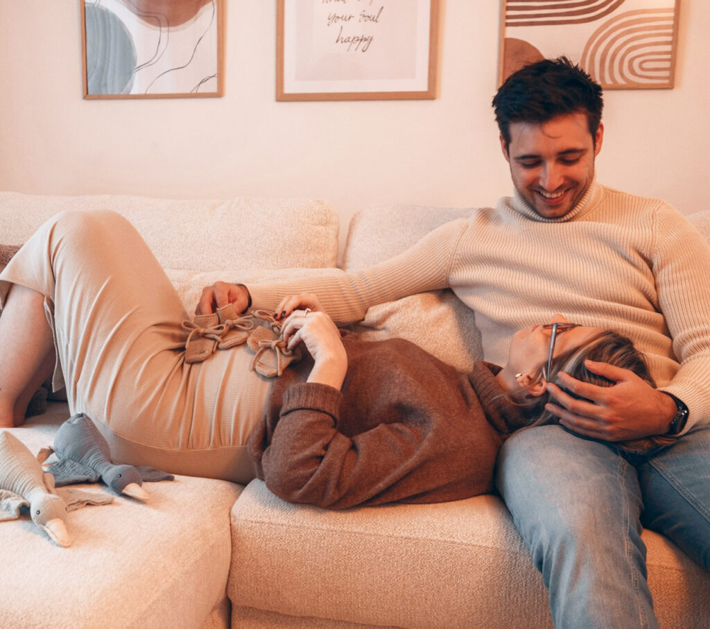 A couple relaxes on a cozy couch, with the pregnant woman lying down with her head on her partner's lap. They are smiling and holding hands, surrounded by soft toys and warm-toned decor, conveying a peaceful, joyful moment of connection during pregnancy.