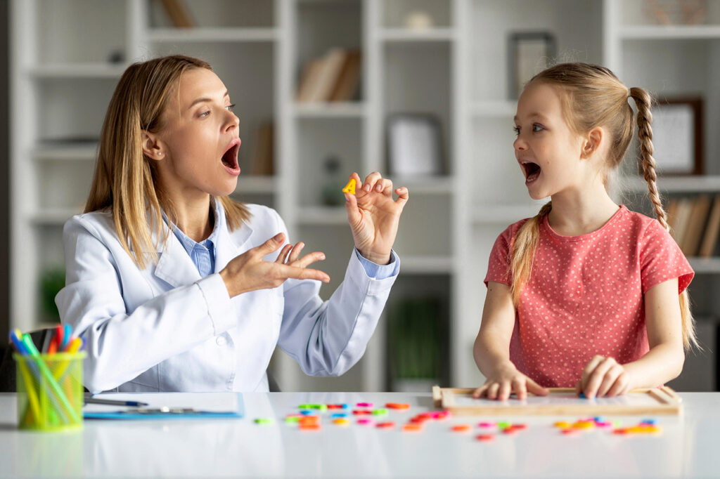 A therapist and a young girl enthusiastically practice speech sounds together using colorful materials—illustrating engaging, play-based speech and language therapy in a supportive environment.
