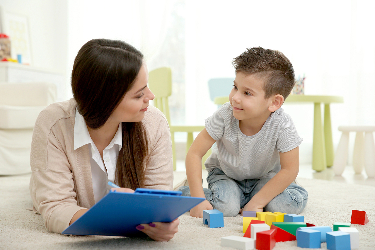 therapist engages with a young boy during a play-based session, using colorful blocks and a clipboard—representing child-centered assessment, support, and therapeutic connection through play.