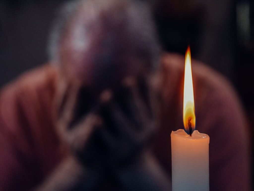 A person holds their head in their hands in a moment of grief or reflection, with a single candle glowing in the foreground—symbolizing loss, healing, and emotional support.