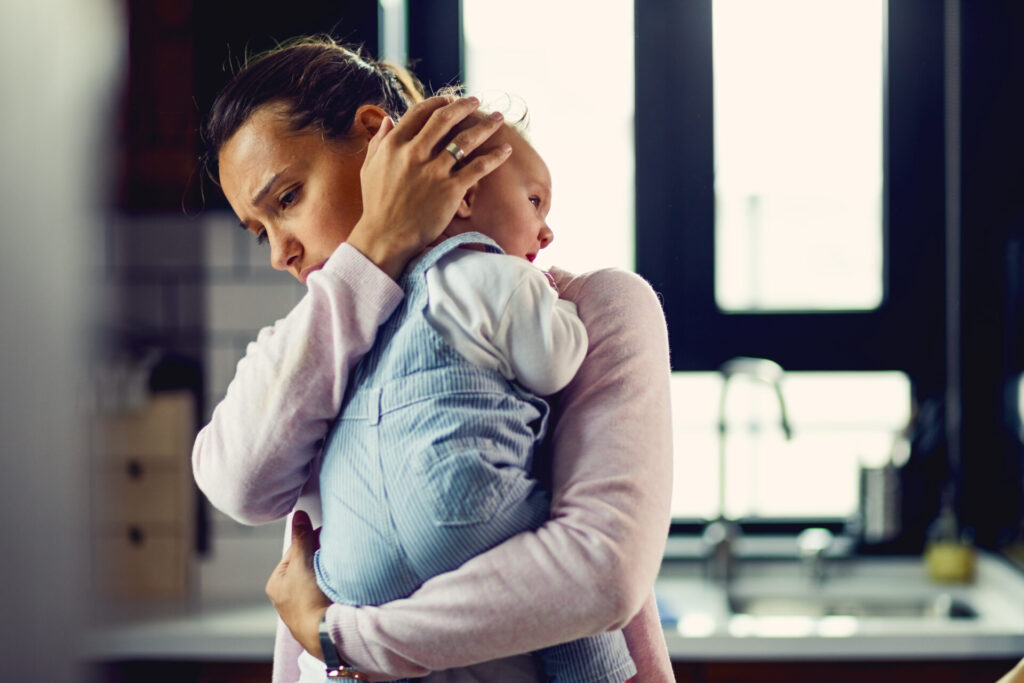 A tired mother holds her baby close in a kitchen, looking overwhelmed, capturing the emotional strain that leads many parents to seek therapy for prenatal depression in Edmonton.